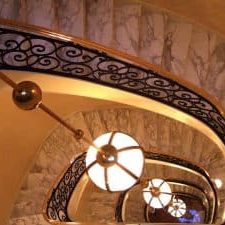 Circular marble stairs with black iron banister with gold trim with large light fixture hanging in the center